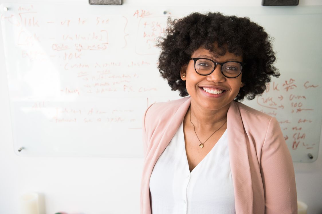 A smiling woman with curly hair and glasses in a pink blazer, head tilted slightly, in front of a whiteboard