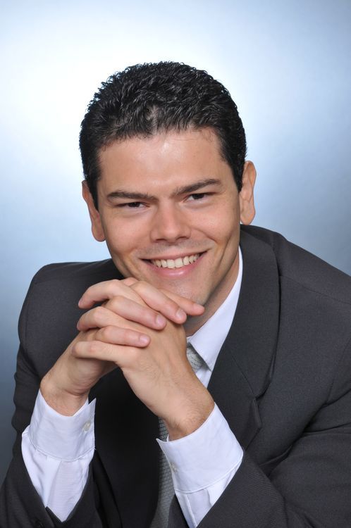 A smiling man in a dark suit leaning toward the camera with his hands clasped together in front of him