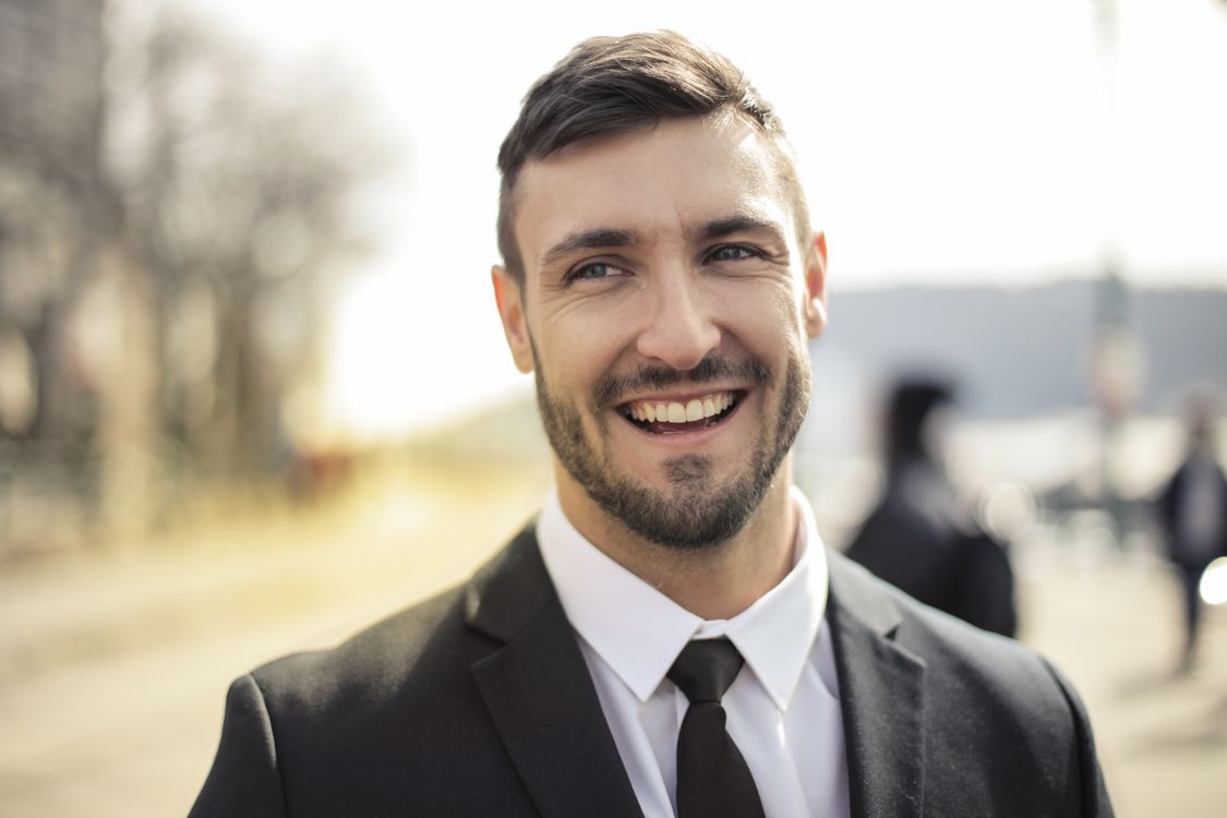 A man in a black suit and tie with a full smile, head slightly tilted, outdoors with a blurred background