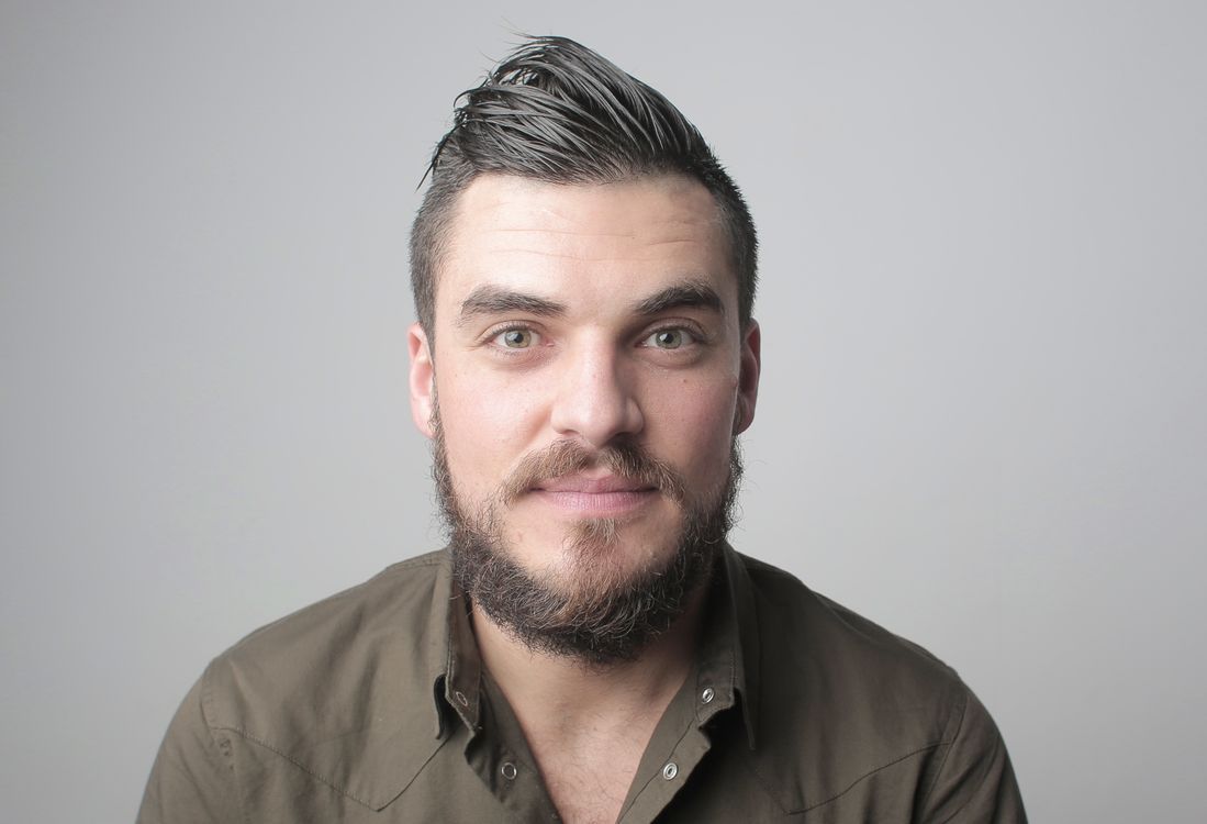 A bearded man with styled hair in an olive shirt, facing the camera with a neutral expression, gray background