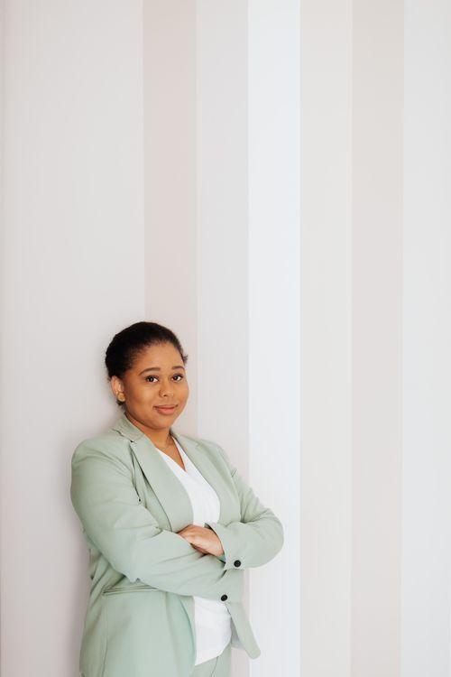 A woman in a pale green suit leaning on a white wall with her arms loosely crossed and a soft smile
