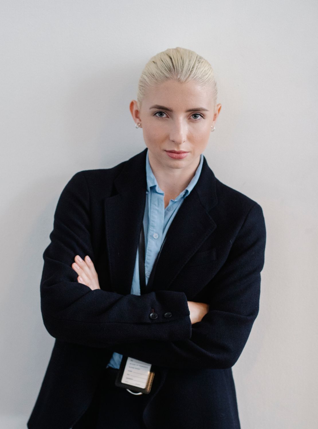 A woman with slicked-back blonde hair in a navy blazer, arms loosely crossed, against a white wall