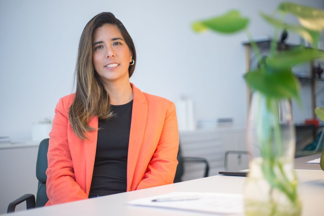 A woman in a coral blazer seated at an office desk with a relaxed, soft smile