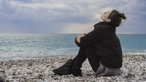 A woman in a black puffer jacket sitting on a pebble beach with her knees up, looking at the sky