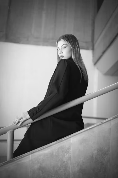 Black and white photo of a woman in a dark coat on a stairway, looking back at the camera over her shoulder
