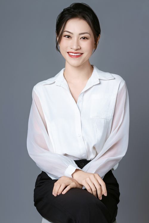 A smiling woman in a white blouse seated and leaning slightly forward with hands on her knees, gray studio